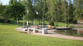 Il Parco Active a Terento L'immagine mostra un parco tranquillo con un piccolo stagno, in cui si trovano sdraio in legno su una superficie sabbiosa. Sullo sfondo ci sono diversi alberi, e l'ambiente è caratterizzato da molta natura e verde.