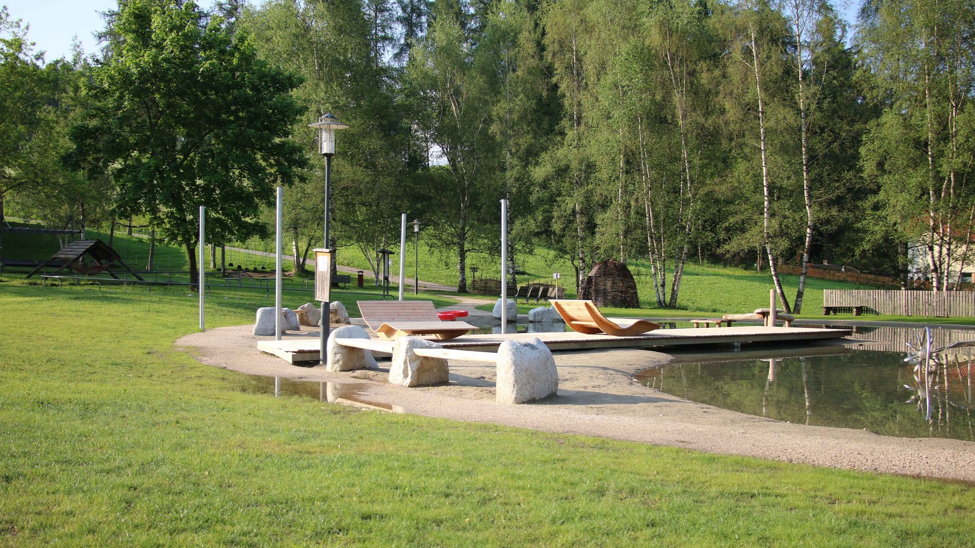Il Parco Active a Terento L'immagine mostra un parco tranquillo con un piccolo stagno, in cui si trovano sdraio in legno su una superficie sabbiosa. Sullo sfondo ci sono diversi alberi, e l'ambiente è caratterizzato da molta natura e verde.