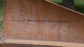 Il Sentiero della Creazione a Rodengo L'immagine mostra una panchina o scultura metallica, sulla quale è inciso il testo "Tag 7 – Vollendung" in diverse lingue. La struttura ha un colore ruggine ed è collocata all'aperto, probabilmente in un ambiente naturale preservato.