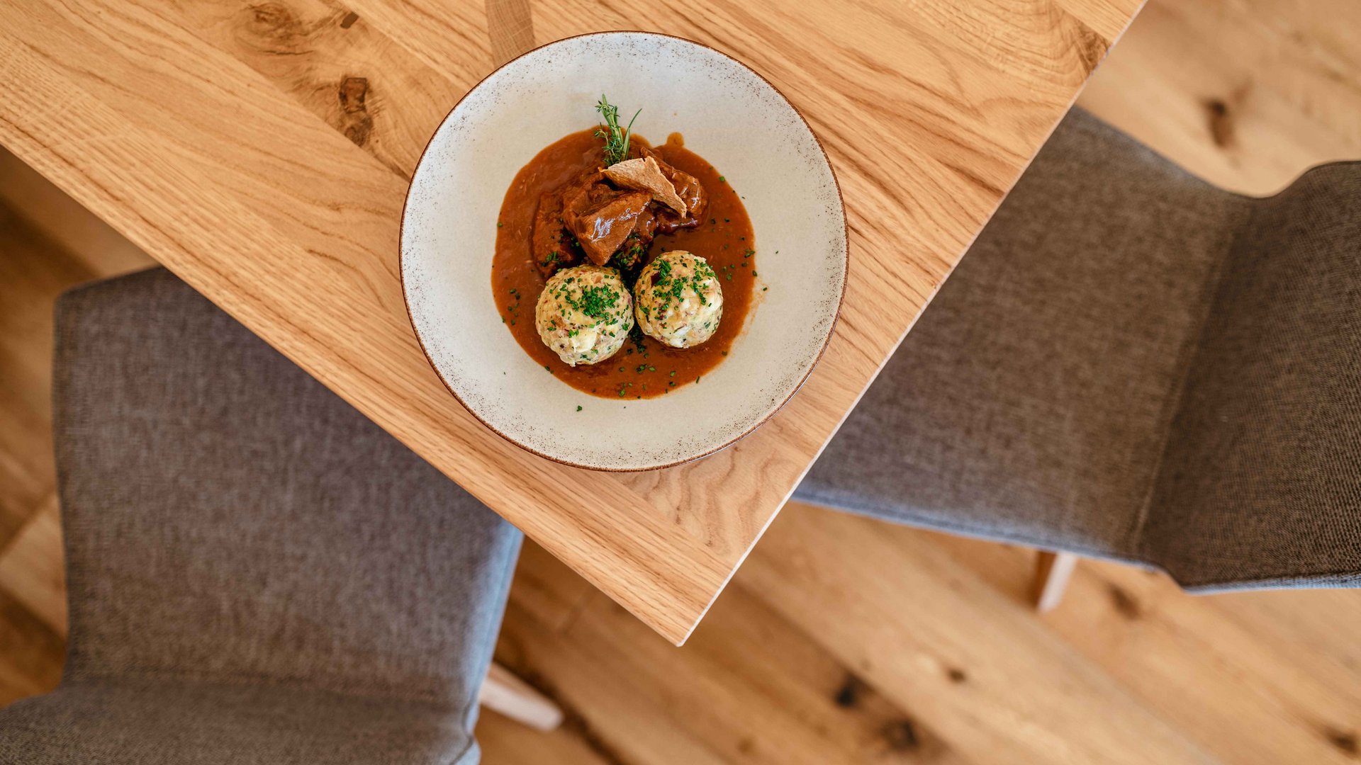 Alla scoperta delle malghe e dei rifugi di Maranza, Valles & Co. Nell'immagine c'è un piatto modernamente impiattato con due canederli di pancetta e gulasch su un tavolo di legno, visto dall'alto, con due sedie vuote sullo sfondo.