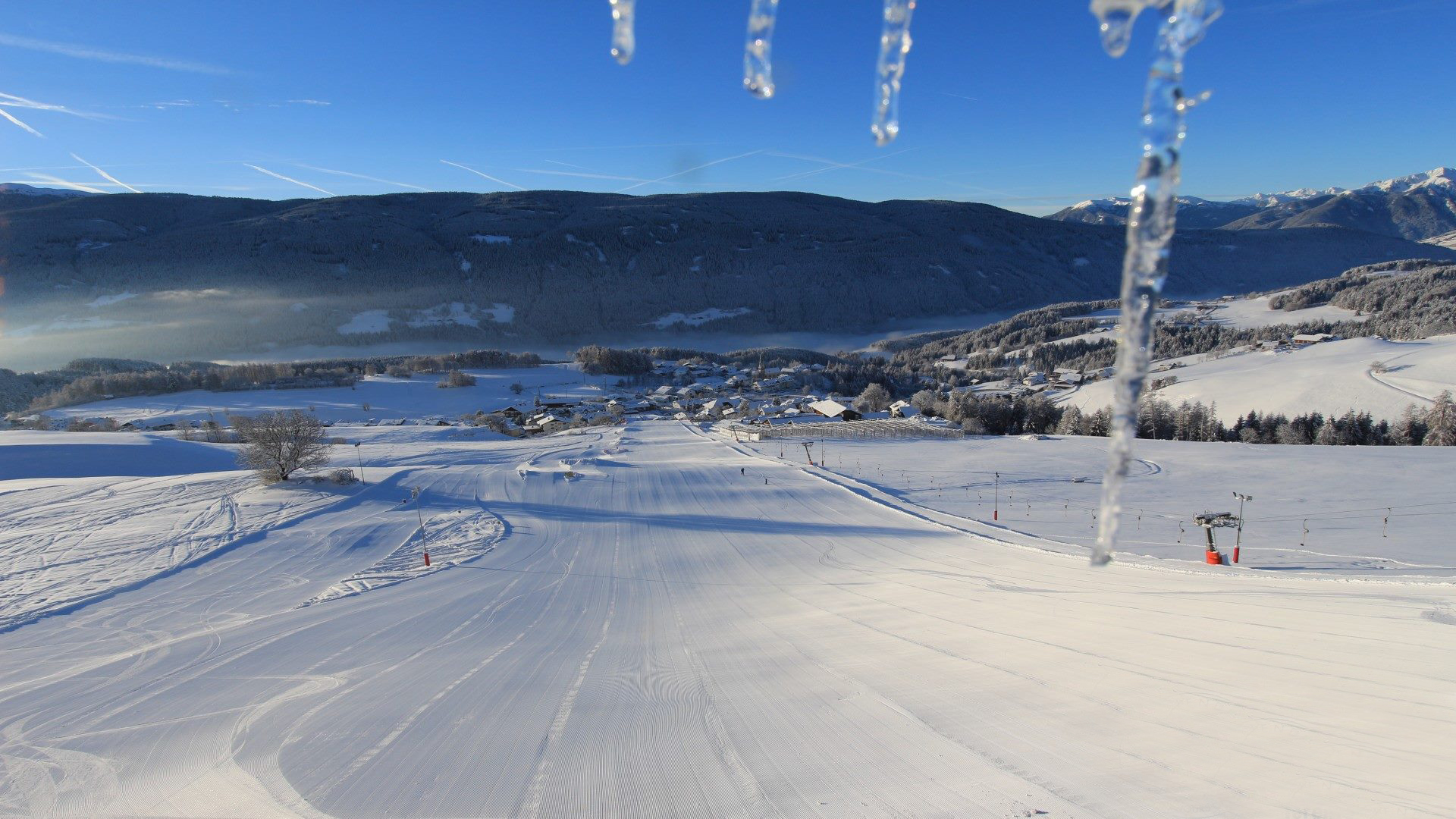 Vacanze sugli sci con i bambini a Terento L'immagine mostra un villaggio innevato con case e una chiesa in mezzo a un paesaggio montano invernale. Sullo sfondo, delle nebbie attraversano la valle, mentre il cielo è chiaro e blu.